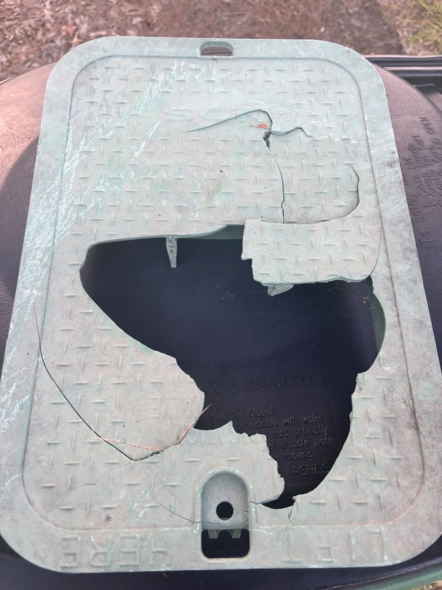 A valve box lid with a large piece broken out of it, sitting in the grass